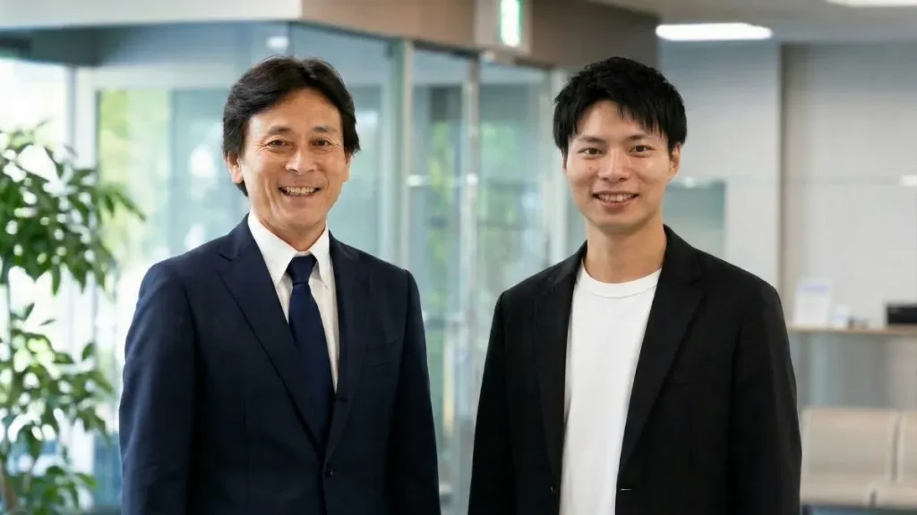 いオフィスで笑顔で並ぶネイビーのスーツ姿の中年男性と黒いジャケット姿の若い男性