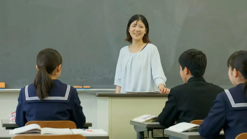 中学校の教室内で、笑顔の女性教師がセーラー服と学ラン姿の生徒たちを前に授業を行う様子